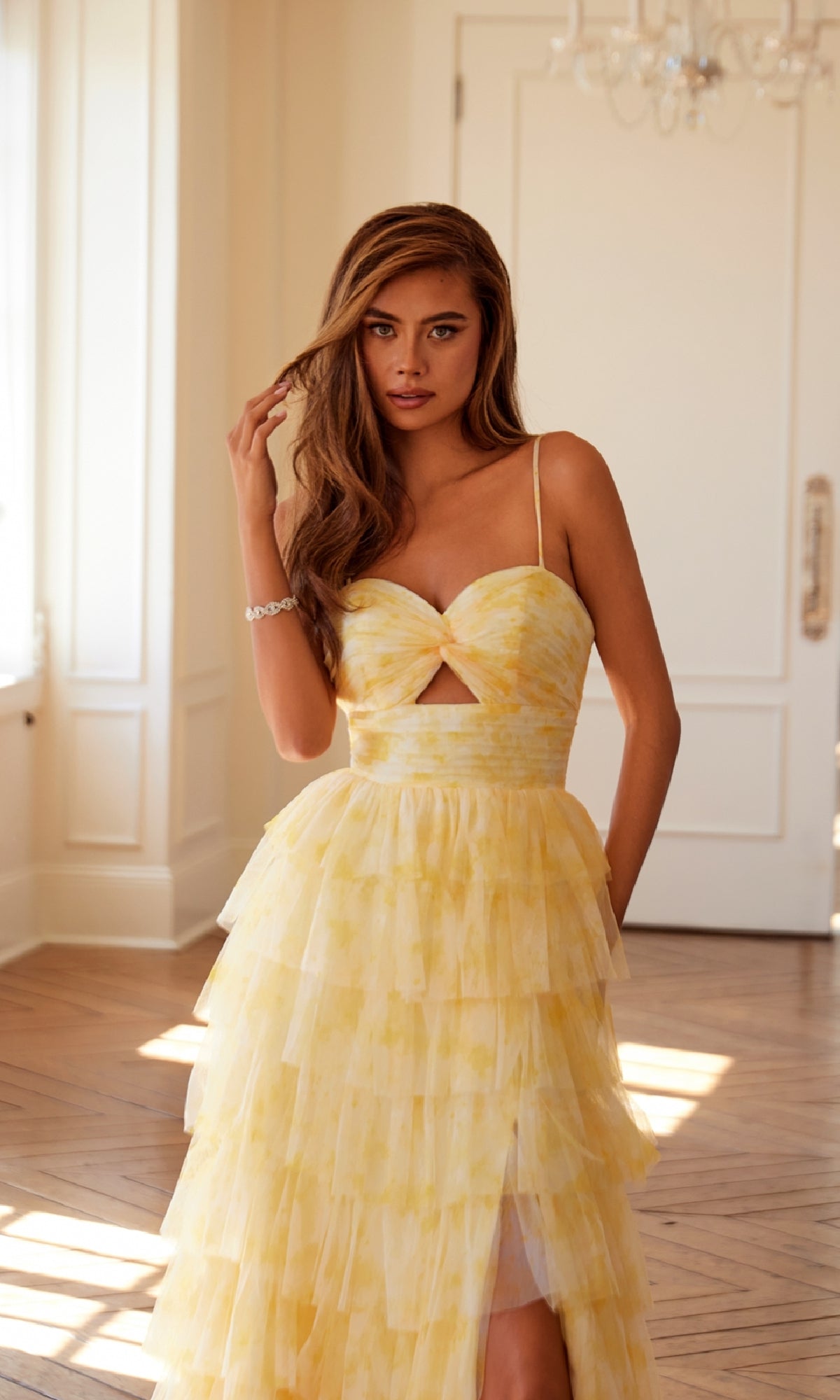 A woman stands indoors in a well-lit room with white walls and wooden floors, wearing the Long Yellow Ruffled Prom Dress: Juliet JT2508A, featuring spaghetti straps and a flowing a-line skirt as she touches her hair.